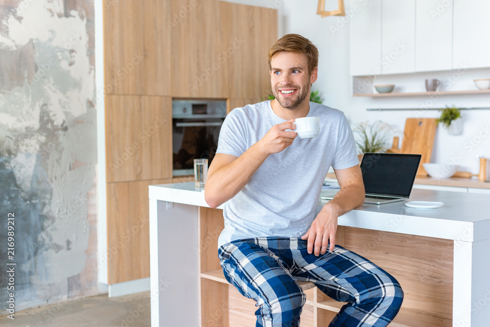 smiling young man drinking coffee at kitchen table with laptop smiling young man drinking coffee at kitchen table with laptop