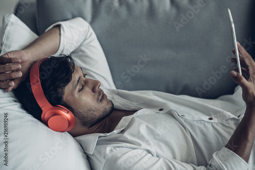 Happy man in headphones with tablet computer relaxing on the sofa in bedroom