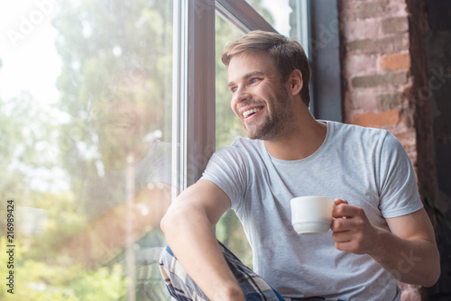 smiling man looking in windows and sitting with cup of coffee on windowsill
