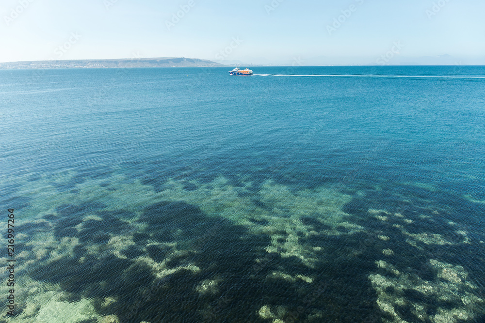 Clear and transparent water of the Mediterranean or Caribbean sea with boats and mountains in the background.