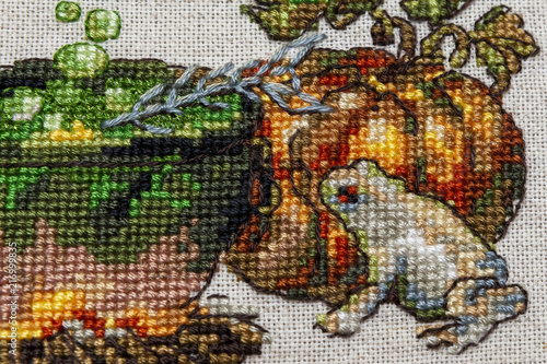 Cross-stitch embroidery with cat in hat, cauldron, toad, bonfire and pumpkin.
