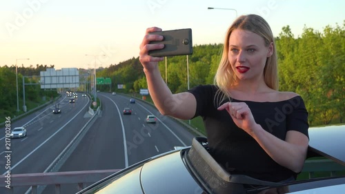 A young beautiful woman stands through a sunroof in a car and takes selfies with a smartphone - a highway in the background