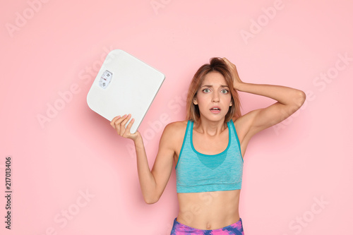 Worried young woman holding bathroom scales on color background. Weight loss diet