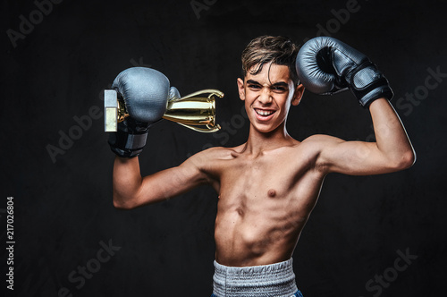 Joyful young shirtless boxer champion wearing gloves holds a winner's cup. Isolated on the dark background. © Fxquadro