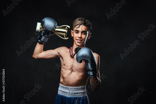 Joyful young shirtless boxer champion wearing gloves holds a winner's cup and the gold medal. Isolated on a dark background. © Fxquadro