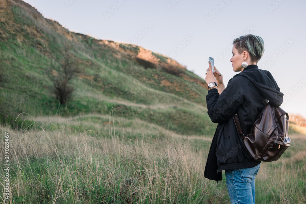 Young woman take a picture on smart phone in the green field with hillon background during spring or autumn time