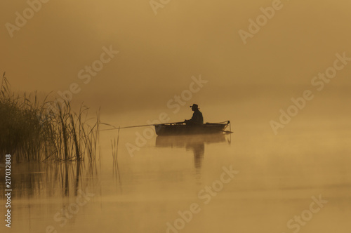 Wędkarz  cieszy się pieknym złotym, mglistym porankiem , łowiąc ryby z małej łodzi, na zalanym słońcem jeziorem