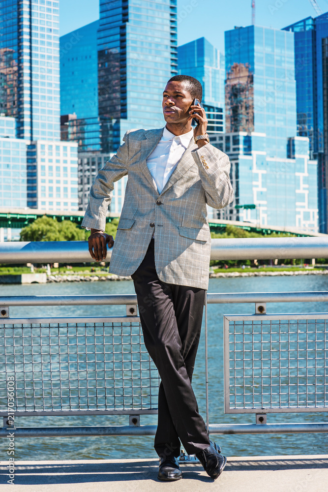 Young African American Businessman traveling in New York City, wearing patterned blazer, white shirt, black pants, leather shoes, standing in business district of Manhattan, talking on cell phone..