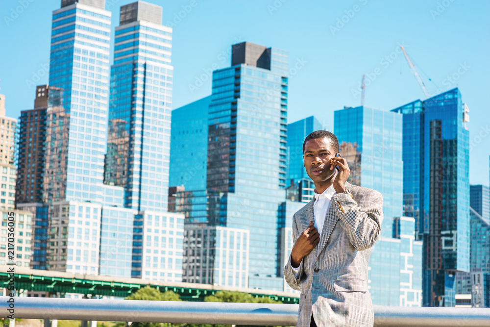 Young African American Businessman traveling in New York City, wearing patterned blazer, white shirt, standing in business district with high buildings of Manhattan under sun, talking on cell phone.. Young African American Businessman traveling in New York City, wearing patterned blazer, white shirt, standing in business district with high buildings of Manhattan under sun, talking on cell phone..