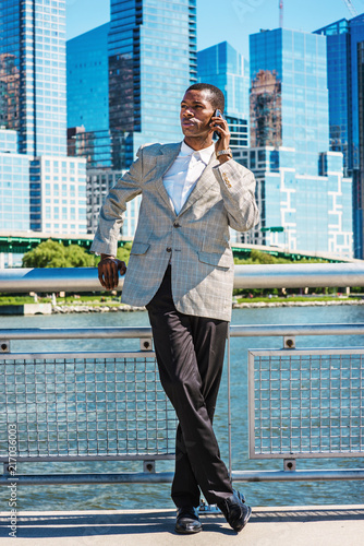 Obraz na płótnie Young African American Businessman traveling in New York City, wearing patterned blazer, white shirt, black pants, leather shoes, standing in business district of Manhattan, talking on cell phone.. w salonie