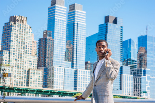 Young African American Businessman traveling in New York City, wearing patterned blazer, white shirt, standing in business district with high buildings of Manhattan under sun, talking on cell phone..