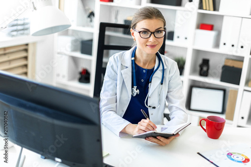 Wallpaper Mural A young girl in a white robe sits at a table in the office and holds a pen and a notebook in her hand. A stethoscope hangs around her neck. Torontodigital.ca