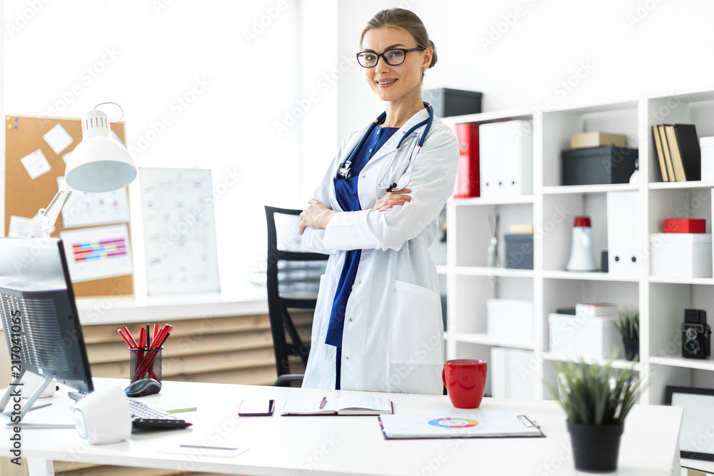 A young girl in a white coat is standing near a table in her office. A stethoscope hangs around her neck.