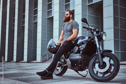 Murais de parede Brutal bearded male in a gray t-shirt and black pants holds a helmet sitting on his custom-made retro motorcycle against skyscraper