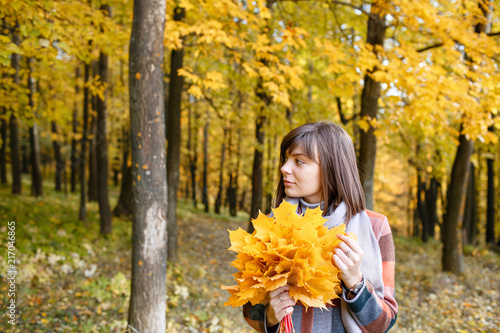 Portrait of young fashion woman outdoor. Brunette woman in autumn park with fashionable plaid coat and scarf. holding yellow leafs