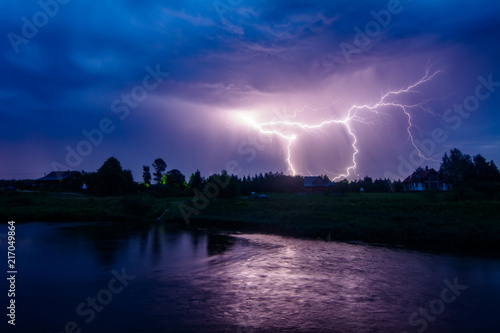 Photo of lightning bolts. Thunderstorm with dramatic clouds. Lots of lightnings over the night village near river bank. Bad weather concept.