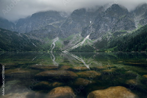 Scenic view of foggy mountains and green forest with a reflection in a lake. Stony shore. Morskie Oko. High Tatras, Zakopane, Poland. Low key view.