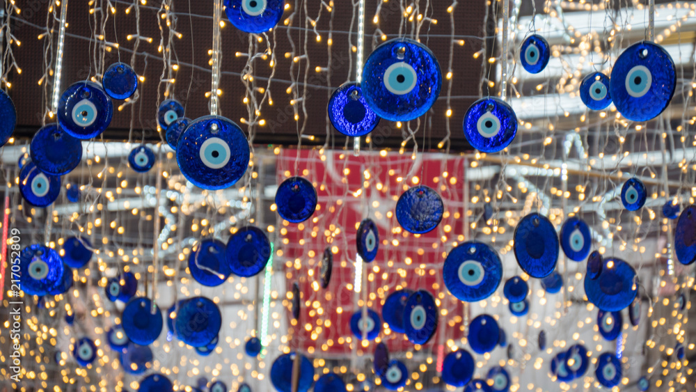 Close up of blue eye-shaped amulets called evil eye or Nazar. Hanged on the roof. Blurred turkish flag and led lights on background. Close up of blue eye-shaped amulets called evil eye or Nazar. Hanged on the roof. Blurred turkish flag and led lights on background.
