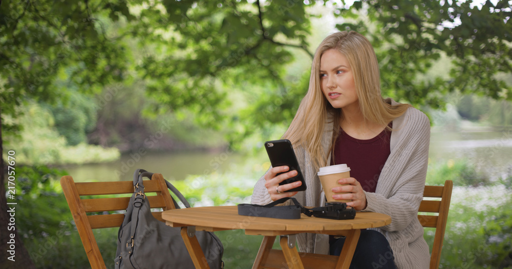 Pretty Caucasian girl drinks coffee and browses web on phone in public park