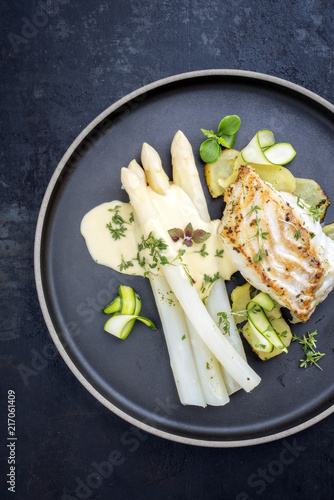 Modern German fried cod fish filet with white asparagus in hollandaise sauce with roast potatoes and sliced zucchini as top view on a plate with copy space