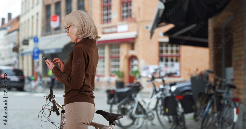 Fototapeta premium Active senior Caucasian woman with bicycle texting on phone on sidewalk