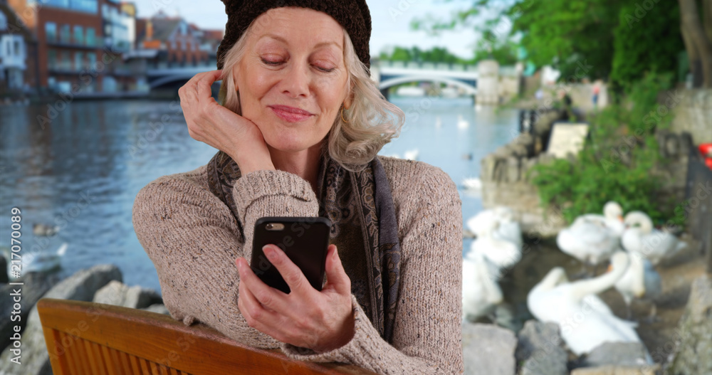 Senior woman on park bench by river in Windsor looking at cell phone with swans