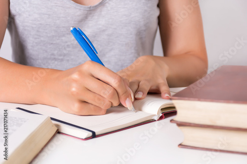 Young girl student writes in notebook with blue pen and full desk with book. Education and back to school concept. Close up, selective focus