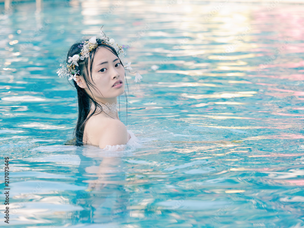 Beautiful fairy in a beautiful white wedding dress and garlands of flowers, standing in swimming pool with sunny ripples on turquoise water, beautiful Chinese girl in water in sunny day. 