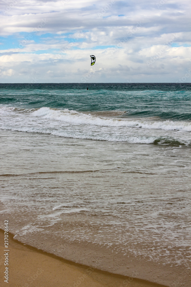 Obraz premium Tide rolling onto the shore with horizon under cloud shadow and kite surfer out in the water - vertical