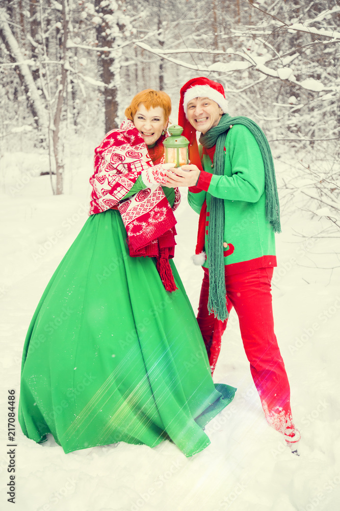 young couple, man and woman, husband and wife are walking in costumes of flowers typical of the elves of Santa's helpers in a winter forest under the snow with a chest full of gifts and a giant candy
