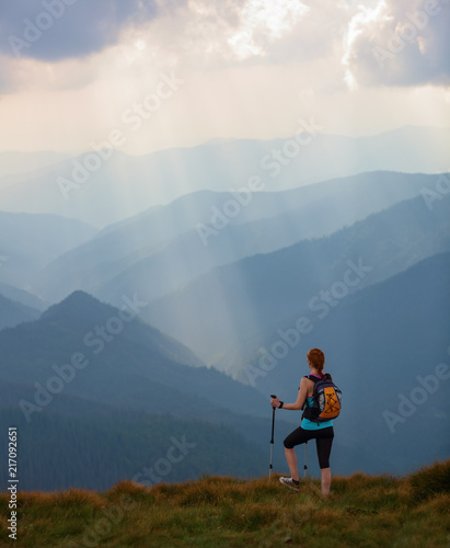 The view with the high mountains in fog. The sun rays are enlighten the forests. The extreme girl with the tracking sticks and back sack is staying on the lawnand watching the nature. Summer scenery.