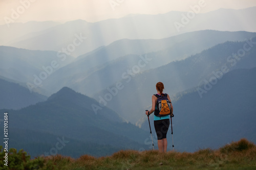 The view with the high mountains in fog. The sun rays are enlighten the forests. The extreme girl with the tracking sticks and back sack is staying on the lawnand watching the nature. Summer scenery. photo