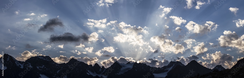 amazing sunset in Tian Shan mountains;  silhouettes of high rocky peaks covered with some snow against blue sky with fluffy clouds illuminated with sun going down; sun rays through the sky; panorama