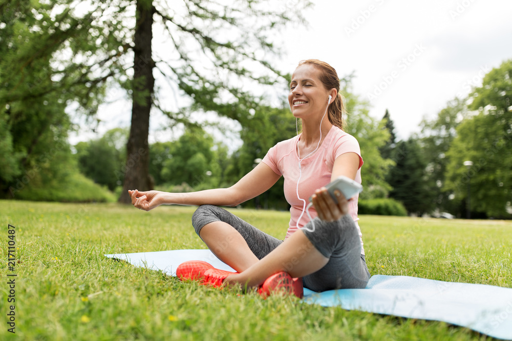 fitness, sport and technology concept - woman with smartphone and earphones meditating on yoga mat at park fitness, sport and technology concept - woman with smartphone and earphones meditating on yoga mat at park