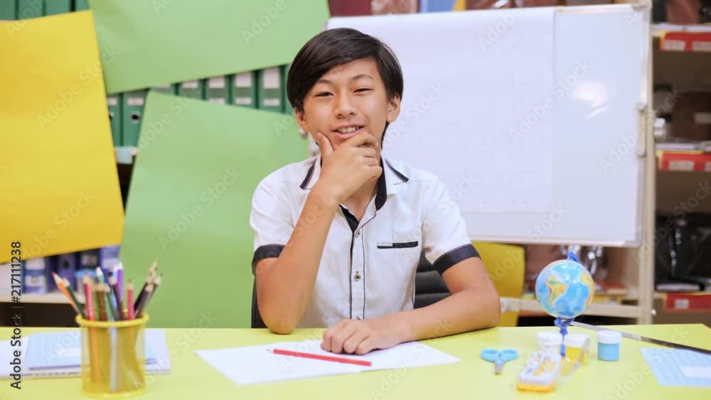 The boy is sitting at the school desk with writing instruments and looks at the camera smiling The boy is sitting at the school desk with writing instruments and looks at the camera smiling