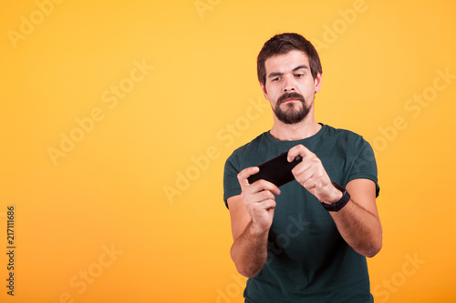 Adult gamer playing video games on his smartphone on yellow background in studio. Leisure activity
