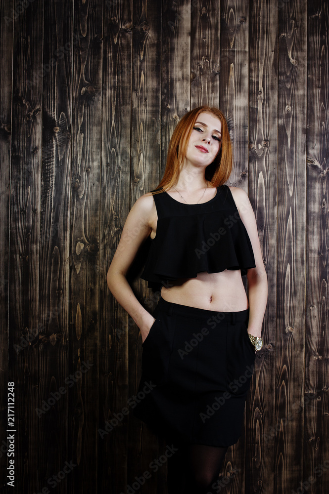 Fototapeta premium Portrait of a beautiful redheaded girl in black top and black skirt posing in the studio next to the wooden wall.