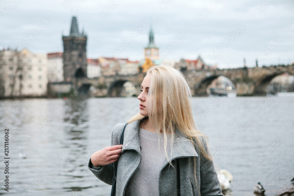 Fashionable blonde with long hair in a coat near Charles Bridge in Prague. Beautiful young woman outdoors.