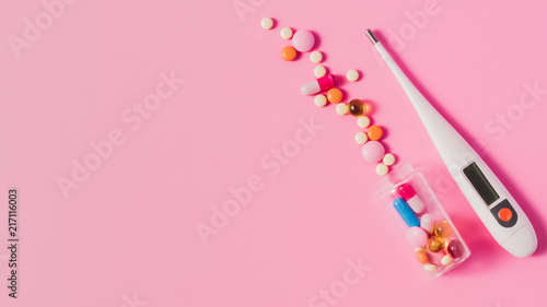 top view of various different pills from bottle and electric thermometer on pink