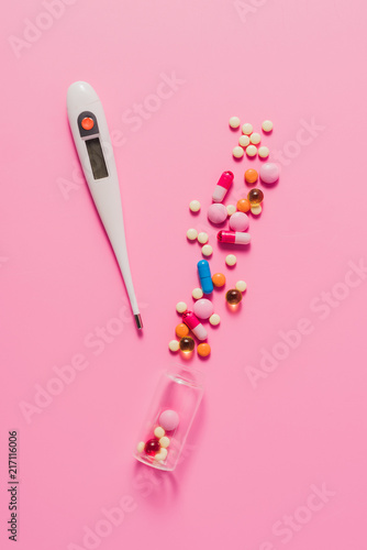 top view of various spilled pills from bottle and electric thermometer on pink