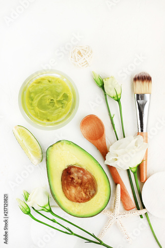 Avocado skin care facial mask in jar, freshly homemade fresh fruit rich in vitamins, lime and flowers for beauty spa treatment. Green color products on white table above background, vertical. 