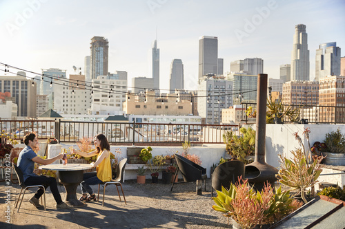 Couple Drinking Wine And Making Toast On Rooftop Terrace With City Skyline In Background