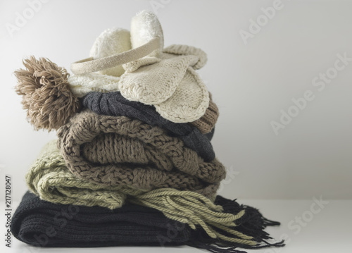 Warm woolen knitted winter and autumn clothes, folded in a pile on a white table. Sweaters, scarves, gloves, hat, headphones. Place for text. Copyspace.