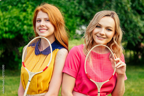 two girlfriends beautiful young ginger redhead irish woman in a yellow dress and european blonde female person in a pink dress holding a tennis badminton racket in the summer park