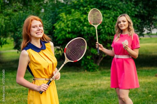 two funny cheerful girlfriends beautiful young ginger redhead irish woman in a yellow dress and european blonde female person in a pink dress playing tennis badminton racket in the summer park