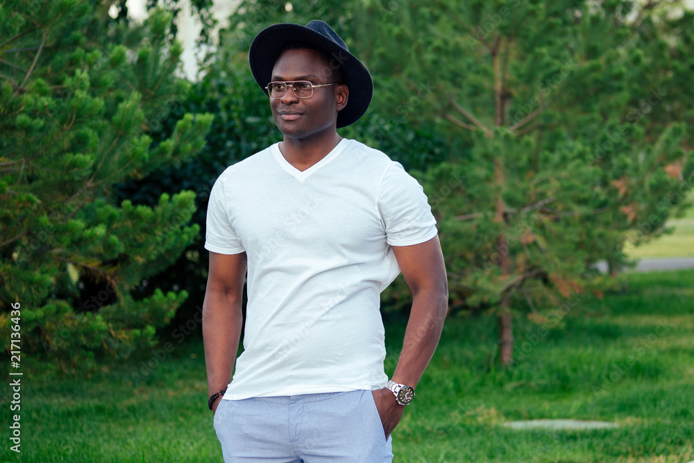 a young and handsome stylish model afro- american man in a stylish suit white t-shirt and a black hat in a summer park . latino american hispanic businessman black guy posing at photoshoot a young and handsome stylish model afro- american man in a stylish suit white t-shirt and a black hat in a summer park . latino american hispanic businessman black guy posing at photoshoot