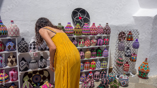 Tourist woman shopping moroccan handcraft souvenirs on the streets of the Medina (Old Town) of a Morocco village. Bright-colored weaved bottles and baskets.