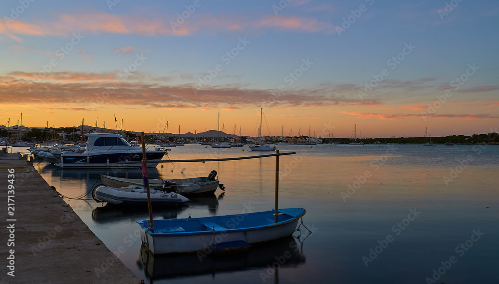 Boats at sunset in mediterranean port under colorful sky and calm waters. Porto Colom, Balearic Islands, Spain