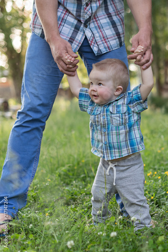 Baby boy holds his father's hands on the village meadow. Father is teaching his son how to walk. © Olga Lyubochkina