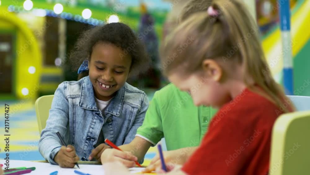 Tracking shot of interested multi ethnic children of primary school age sitting at table in play center and drawing picture together with colored felt tip pens Tracking shot of interested multi ethnic children of primary school age sitting at table in play center and drawing picture together with colored felt tip pens